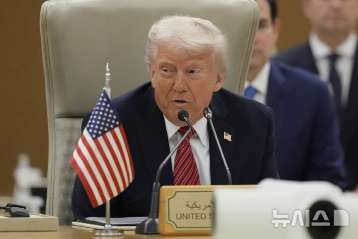 정장 차림의 남성이 미국 국기와 'UNITED STATES'라고 적힌 명패 앞에 앉아 마이크로 발언하고 있다. 회의실 분위기이며 주변에 다른 사람들이 보인다.