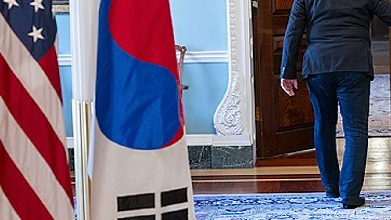 미국 국기와 대한민국 국기가 나란히 서 있고, 한 사람이 문을 열고 방을 나가고 있다.