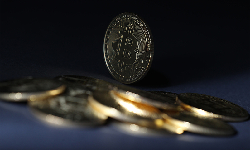 비트코인 로고가 새겨진 동전 여러 개가 어두운 배경 위에 놓여 있고, 한 개의 동전이 세워져 있다.