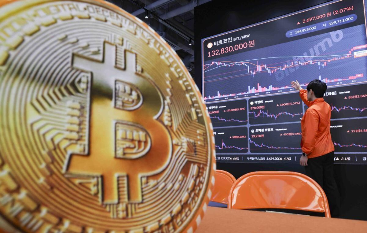 비트코인 로고가 크게 보이고, 뒤에는 비트코인 시세와 차트가 표시된 대형 전광판 앞에서 한 남성이 차트를 가리키고 있다.