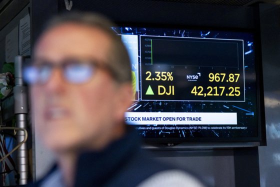 주식 시장의 거래가 시작되었음을 알리는 전광판이 보이고, 전광판에는 다우존스 지수가 2.35% 상승해 42,217.25를 기록하고 있다는 정보가 표시되어 있다. 화면 앞에는 흐릿하게 한 남성이 보인다.