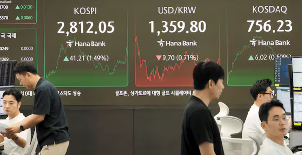 여러 명의 사람들이 일하고 있는 증권사 사무실 내부에서, 대형 전광판에 KOSPI, USD/KRW, KOSDAQ 지수와 변동 그래프가 표시되어 있다.