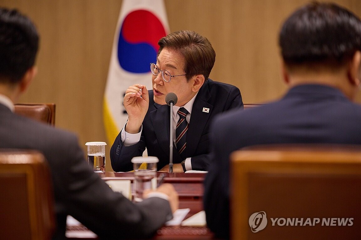 정장 차림의 남성이 마이크 앞에서 회의 중 발언하고 있으며, 뒤에는 태극기가 보인다. 주변에 다른 사람들이 함께 앉아 있다.