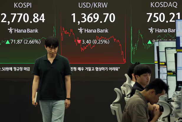 증권사 트레이딩룸에서 여러 명의 직원들이 컴퓨터 앞에 앉아 일하고 있고, 뒤에는 KOSPI, USD/KRW, KOSDAQ 지수가 표시된 대형 전광판이 보인다.