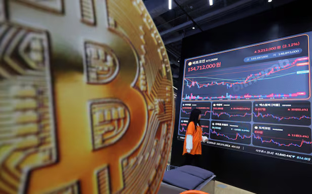 비트코인 로고가 크게 보이고, 한 여성이 암호화폐 시세 차트가 표시된 대형 전광판을 바라보고 있다.