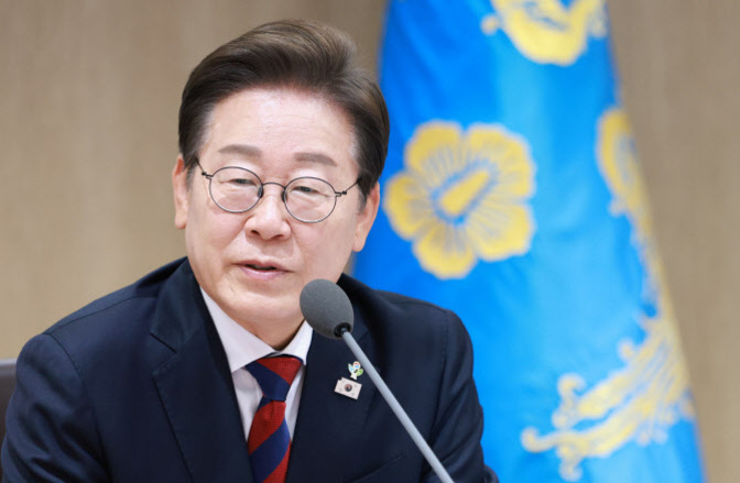 정장을 입은 남성이 안경을 쓰고 마이크 앞에서 말하고 있으며, 뒤에는 파란색 바탕에 노란 무늬가 있는 깃발이 있다.